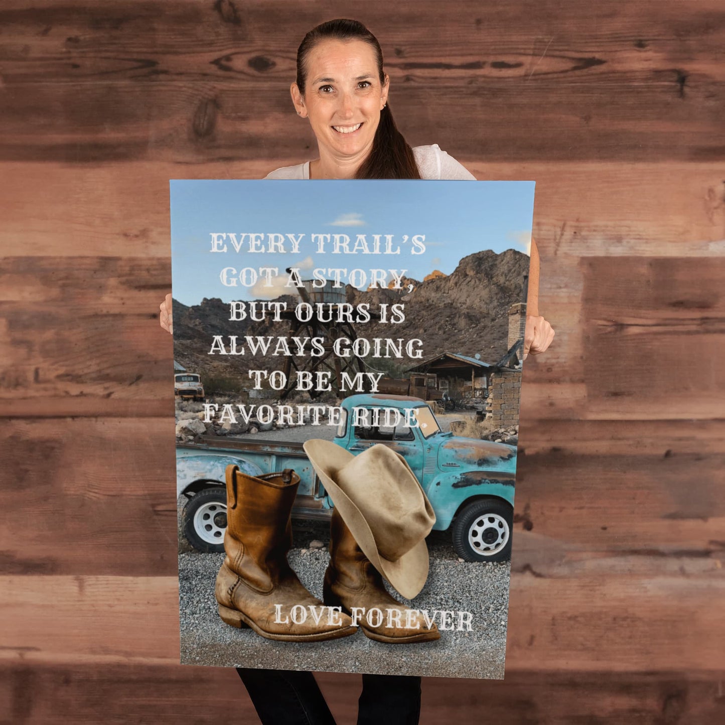 Person holding a poster with a scenic design featuring a blue truck, cowboy boots, and a hat against a wooden background.